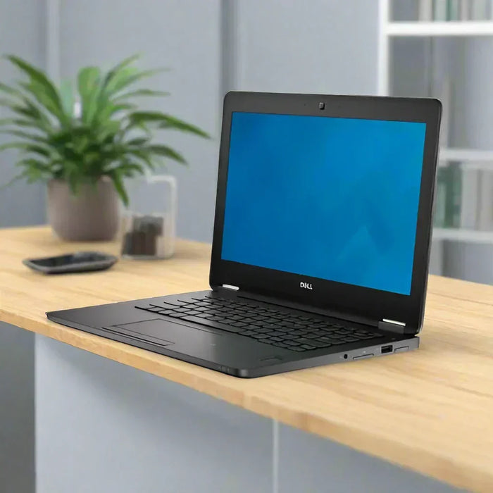A certified refurbished Dell Latitude E7270 i7 6600U 2.6GHz 8GB 256GB SSD 12.5" W10P laptop sits open on a light wooden desk with a blue screen, next to a green potted plant, remote control, and background organizer.