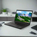 A refurbished black Lenovo ThinkPad T450s i7 sits open on a white desk, displaying a waterfall screensaver. Nearby are a smartphone, notebook, and keyboard, with a blurred office in the background.