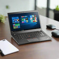 A certified refurbished Lenovo ThinkPad T460s i7 6600U 2.6GHz 8GB 256GB SSD W10P 14 Laptop sits on a wooden desk displaying the Windows 10 start menu, with a wireless mouse, mouse pad, and clipboard nearby; background is softly blurred.