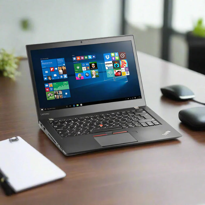 A certified refurbished Lenovo ThinkPad T460s i7 6600U 2.6GHz 8GB 256GB SSD W10P 14 Laptop sits on a wooden desk displaying the Windows 10 start menu, with a wireless mouse, mouse pad, and clipboard nearby; background is softly blurred.