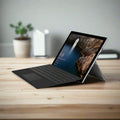 A refurbished Microsoft Surface Pro 4 1724 i5 6300U 2.4GHz 8GB 256GB 12" Touch W10P sits on a wooden desk with its detachable keyboard and stand, displaying a nature wallpaper, with blurred plants and office items in the background.
